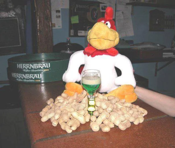 Un gallo di peluche appoggiato sul bancone dietro una montagna di noccioline, con accanto un bicchierino di liquore verde