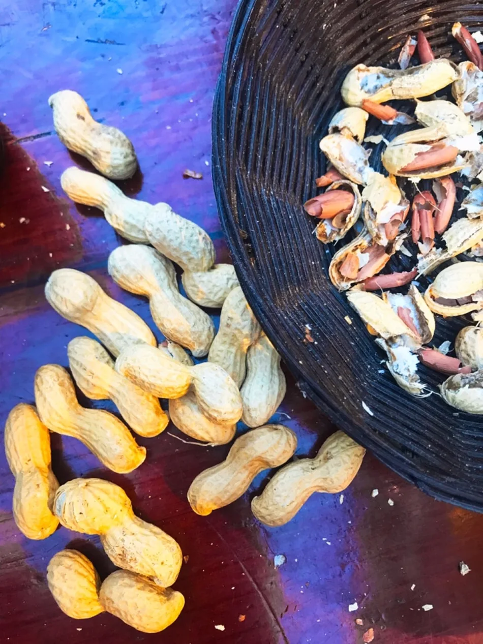 Noccioline con il guscio sparse su un tavolo di legno dipinto di viola, accanto a una ciotola di vimini piena di gusci rotti